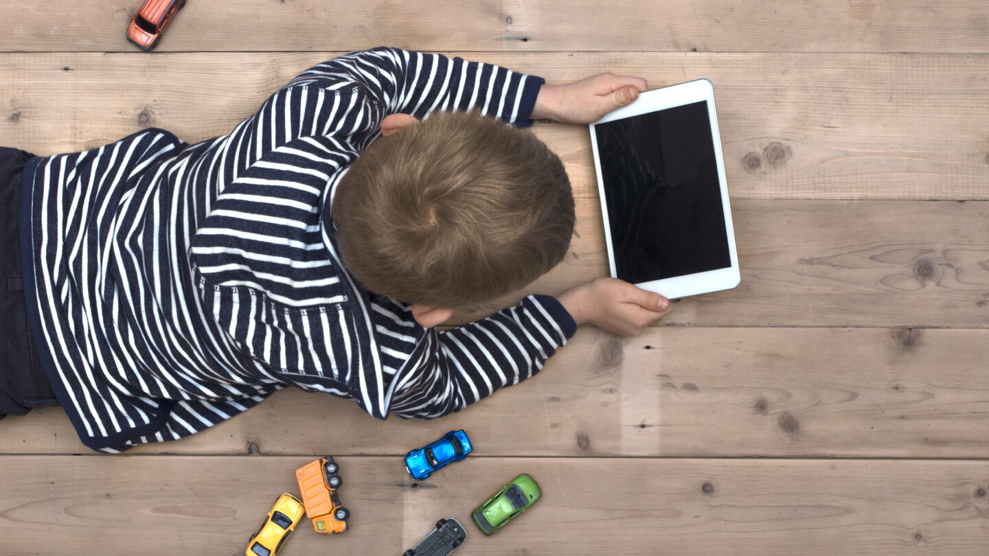 Photo of Top view header image with child laying down on the floor watching a movie on his tablet instead of playing with his toy cars