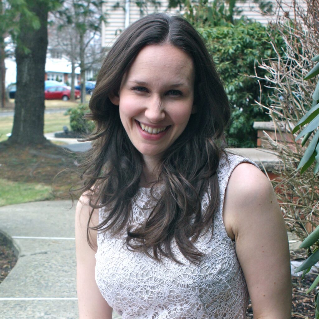 A woman with long brown hair, wearing a sleeveless white lace top, smiles while standing outdoors on a path with trees and shrubs in the background.