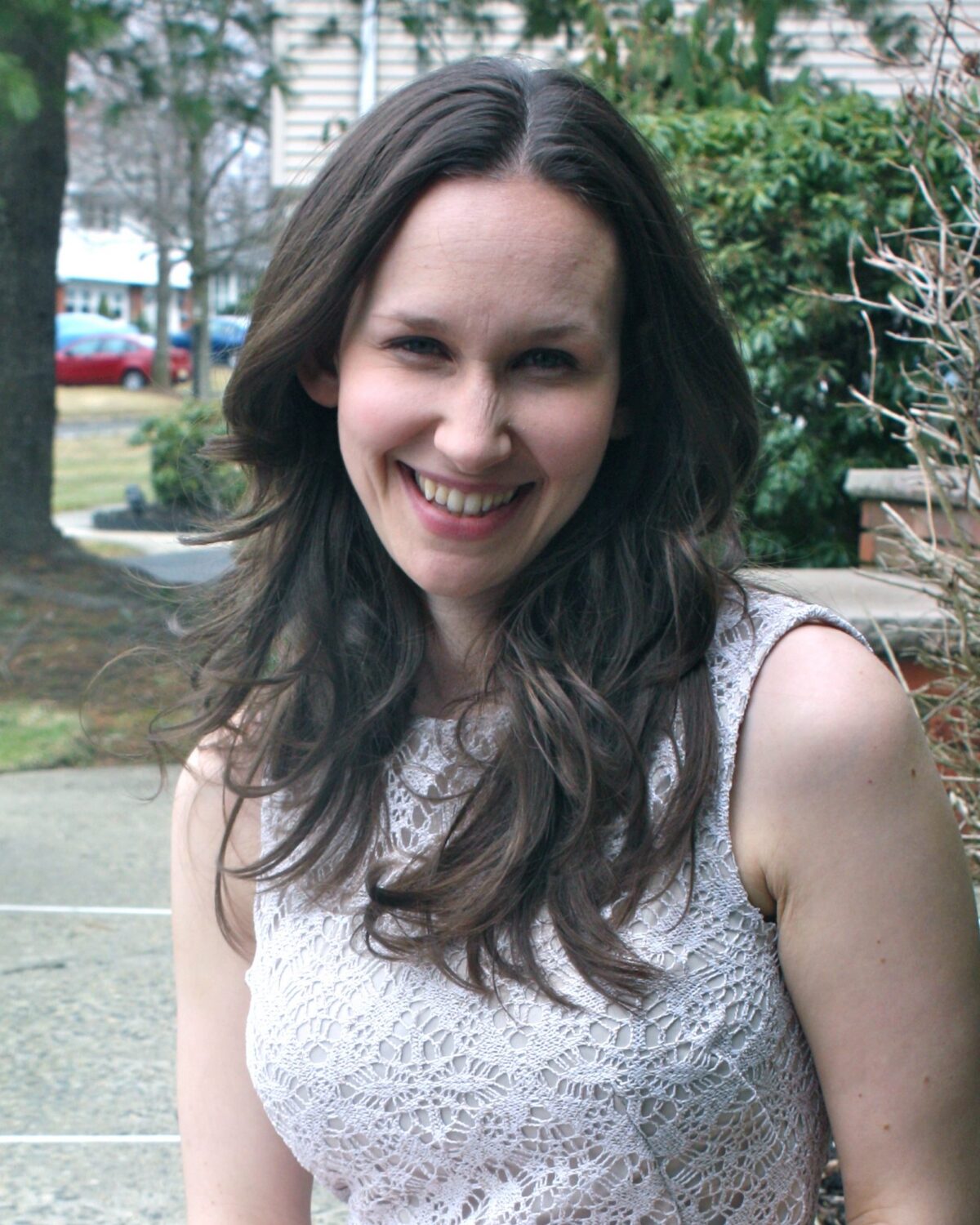 A woman with long brown hair, wearing a sleeveless white lace top, smiles while standing outdoors on a path with trees and shrubs in the background.