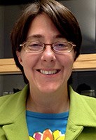 A person with short brown hair and glasses, wearing a green jacket and blue shirt, smiling in an indoor setting.