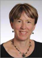 A woman with short brown hair, wearing earrings and a beaded necklace, smiles at the camera against a light gray background.