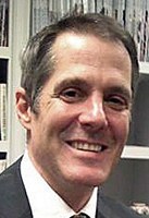 A man in a suit smiling, with short brown hair, in front of shelves filled with books.