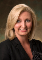 A woman with shoulder-length blonde hair, wearing a black blazer, smiles at the camera in a professional portrait with a brown and green background.