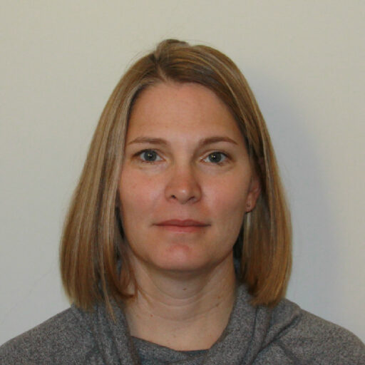 A woman with straight, shoulder-length blonde hair and fair skin looks at the camera with a neutral expression. She is wearing a grey top and is in front of a plain light background.