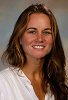 A woman with long brown hair, wearing a white shirt, smiles at the camera against a blurred, neutral background.