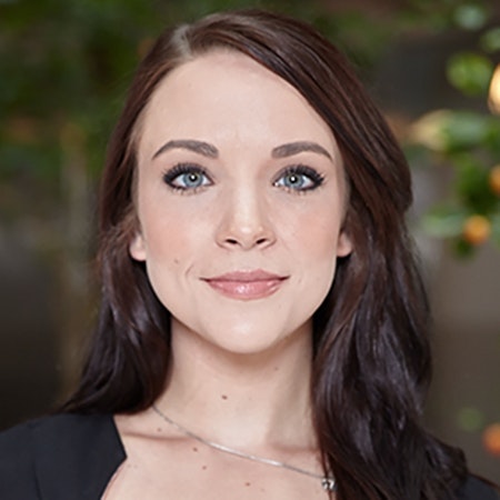 A woman with long dark brown hair and blue eyes, wearing a black top and a necklace, stands in front of a blurred green background.