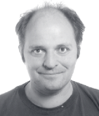 A man with a receding hairline smiles at the camera. He has short hair on the sides and is wearing a dark shirt. The photo is black and white with a plain background.