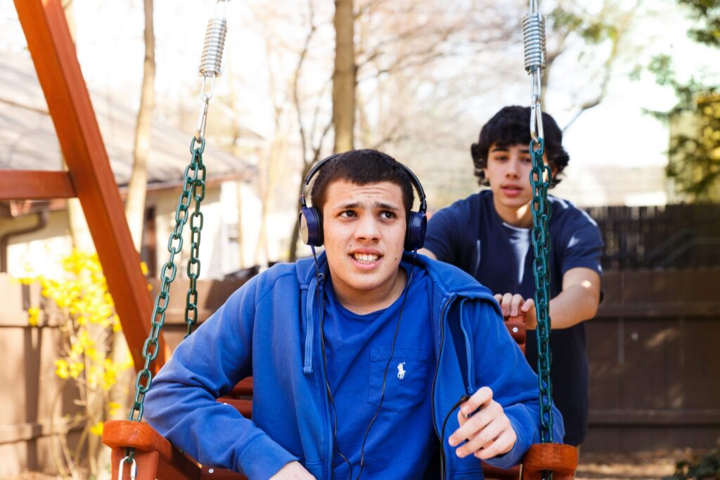A young man wearing headphones sits on a swing while another young man stands behind him, seemingly pushing the swing, both outdoors on a sunny day.