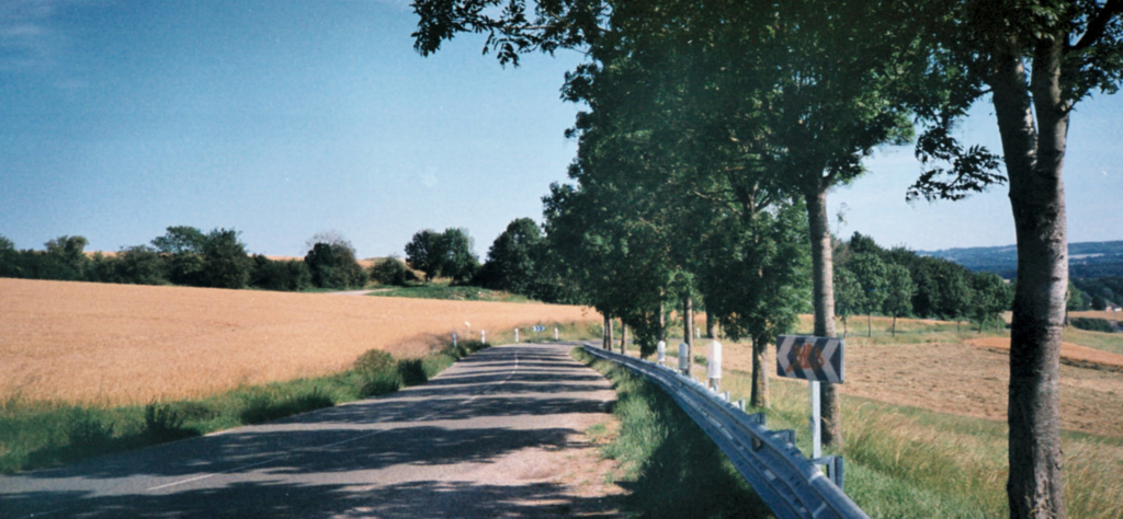 A rural road curves through fields and trees under a clear blue sky, with a metal guardrail and a chevron road sign visible on the right side.
