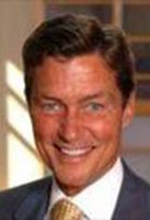 A man wearing a suit and tie is smiling at the camera. The background is blurred with some light and shadow patterns.