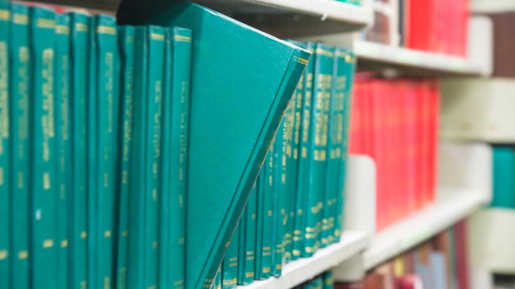 A row of teal hardcover books on a white bookshelf, with one book partially pulled out at an angle. Red books are visible on shelves in the background.