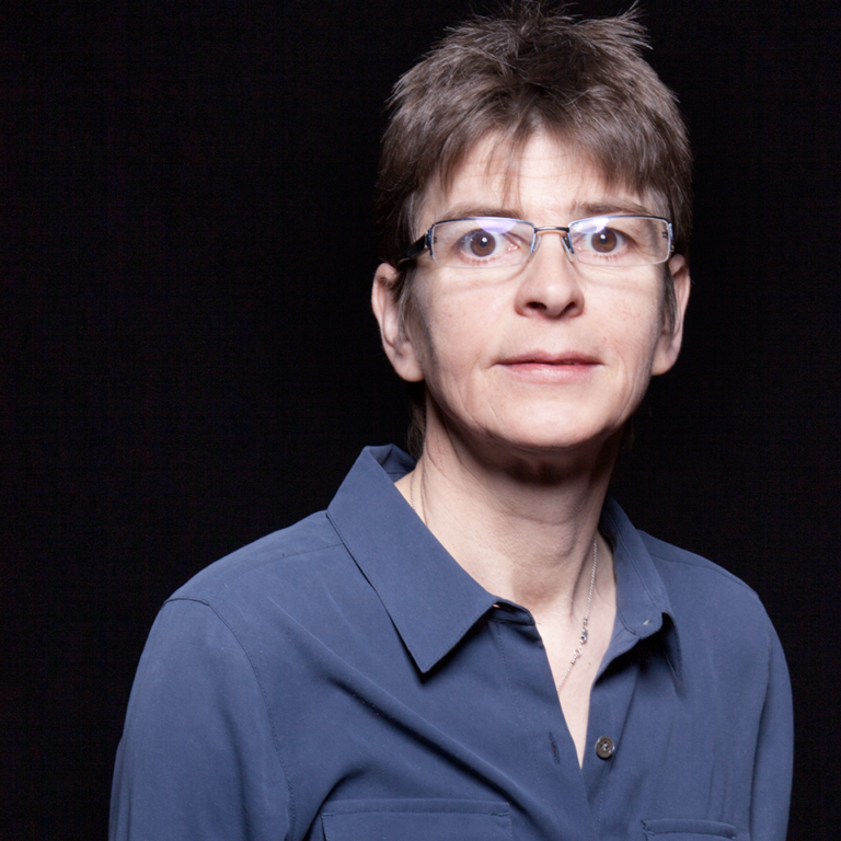 A person with short brown hair and glasses, wearing a blue button-up shirt, stands against a plain black background, looking toward the camera with a neutral expression.