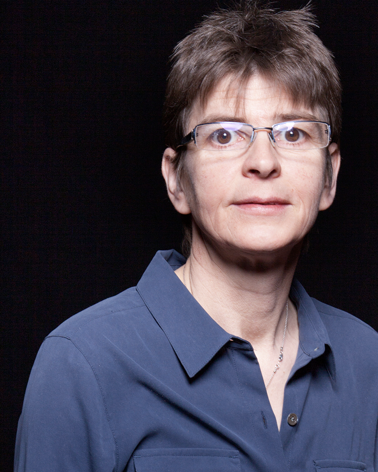 A person with short brown hair and glasses, wearing a blue button-up shirt, stands against a plain black background, looking toward the camera with a neutral expression.
