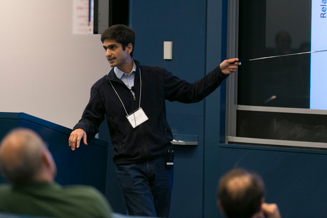 Michael Desai leaning on a lectern while using a metal stick to point to a slideshow