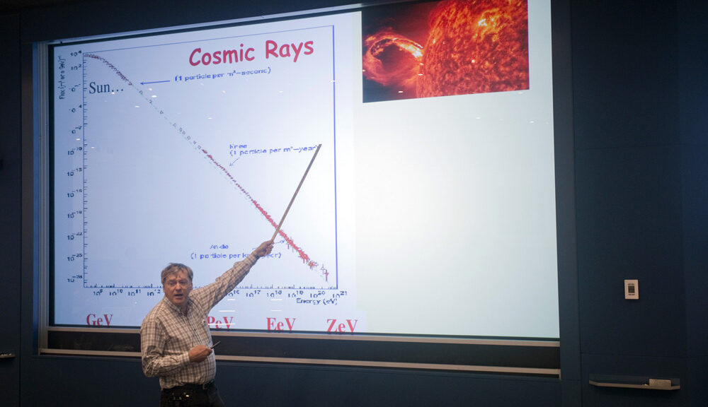 Man pointing to a projected slideshow titled Cosmic Rays