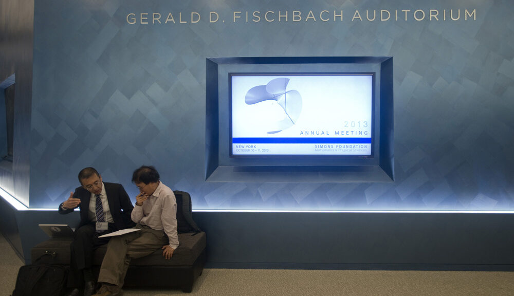 Two people sitting by a wall with 3D lettering that says Gerald D. Fischbach Auditorium