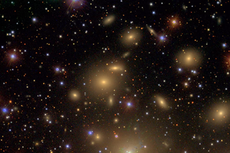 A dense cluster of galaxies glowing yellow-orange among countless stars and distant galaxies scattered across the blackness of space.