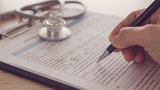 A person fills out a medical form on a clipboard with a pen, while a stethoscope lies on the table nearby.