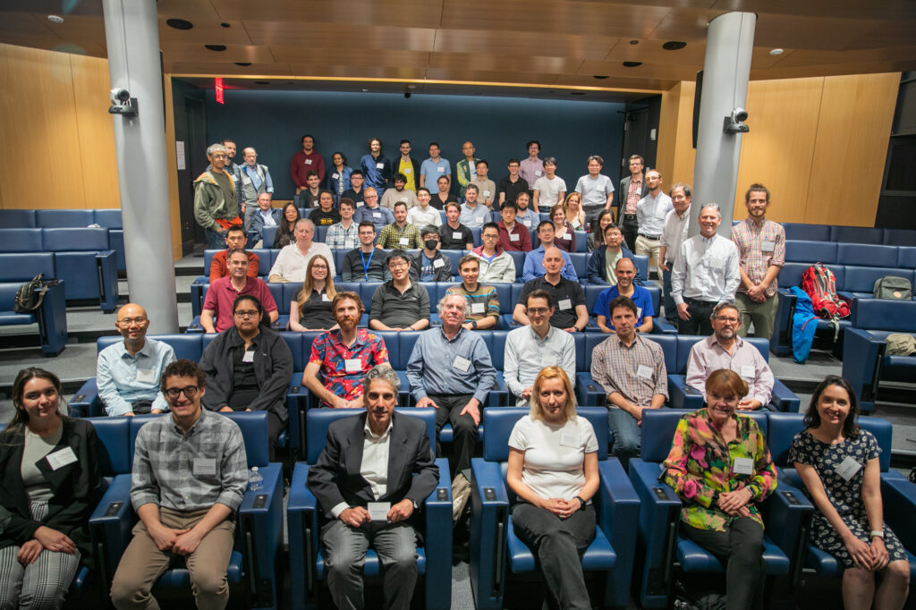 A large group of people sits and stands in rows inside a modern lecture hall, facing the camera, with blue seats and bright lighting.
