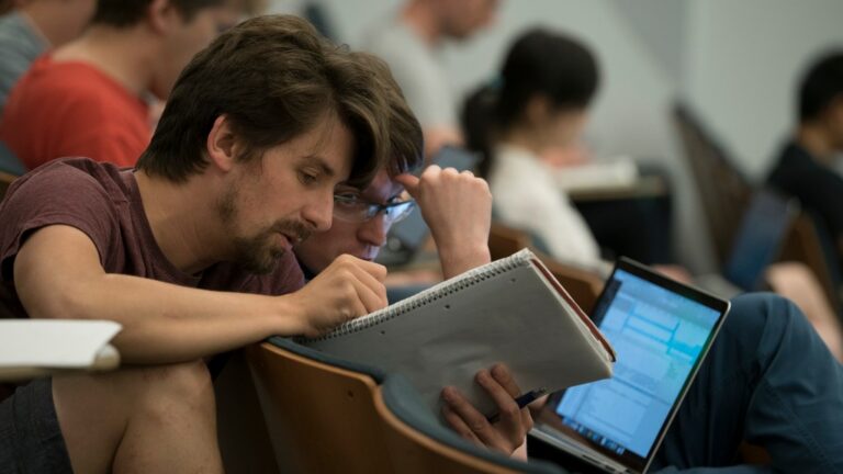 Two men sitting in different lecture hall rows, collaborating on a notebook