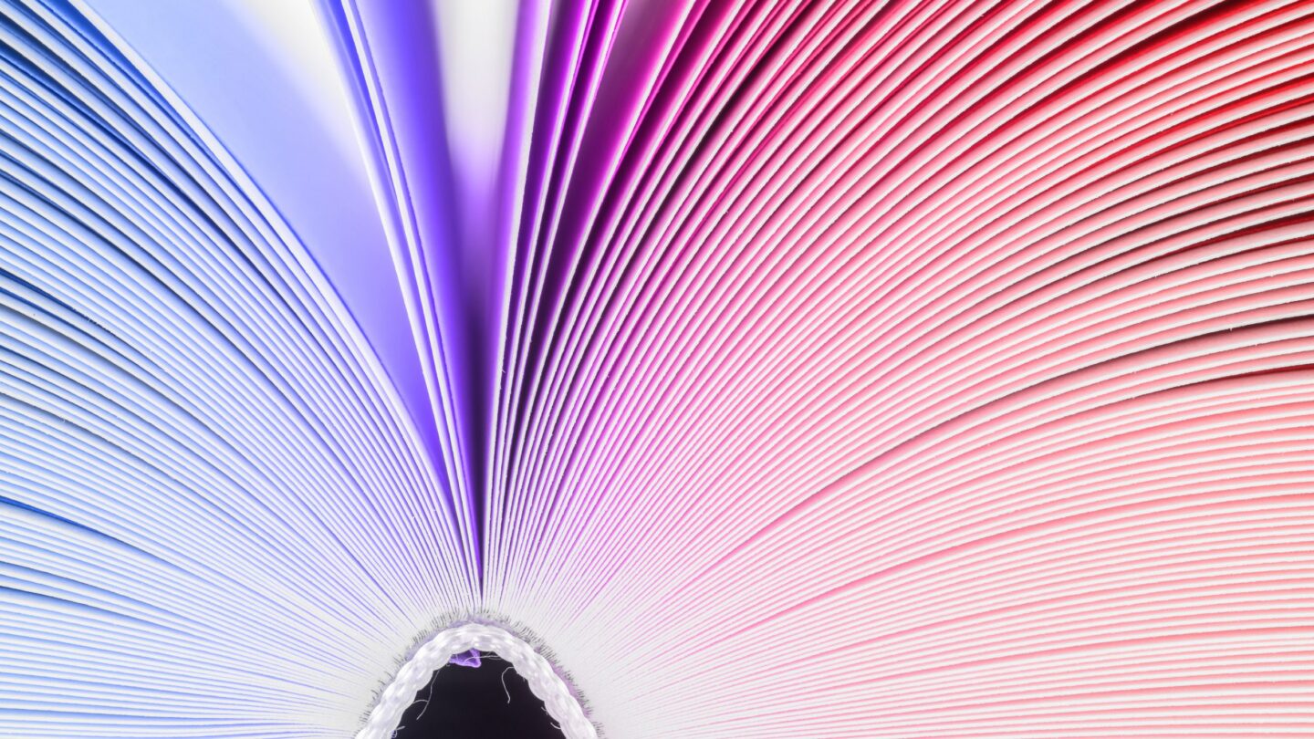 Close-up of an open book viewed from the bottom, with pages fanned out. The left side is tinted purple, transitioning to pink and red on the right, creating a vibrant, abstract pattern.