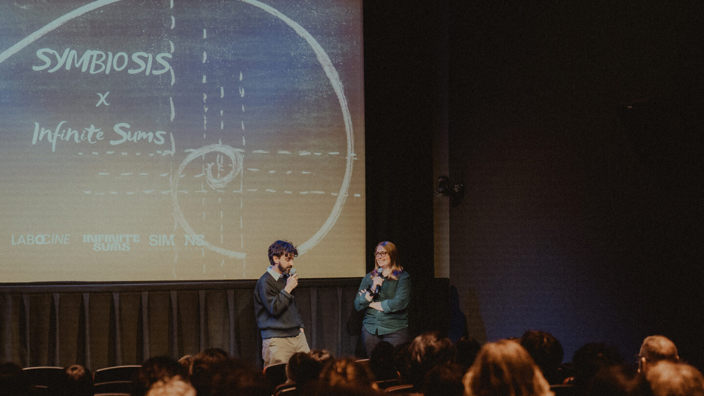 A photo of Sonya Hansen and Diego Murillo at the Symbiosis film screening, standing in front of a screen that says Symbiosis x Infinite Sums.