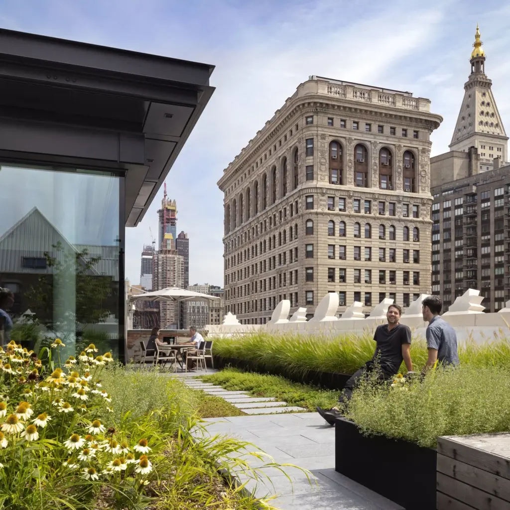 Three people walk and talk on a rooftop garden with greenery and flowers, surrounded by tall historic buildings under a blue sky.