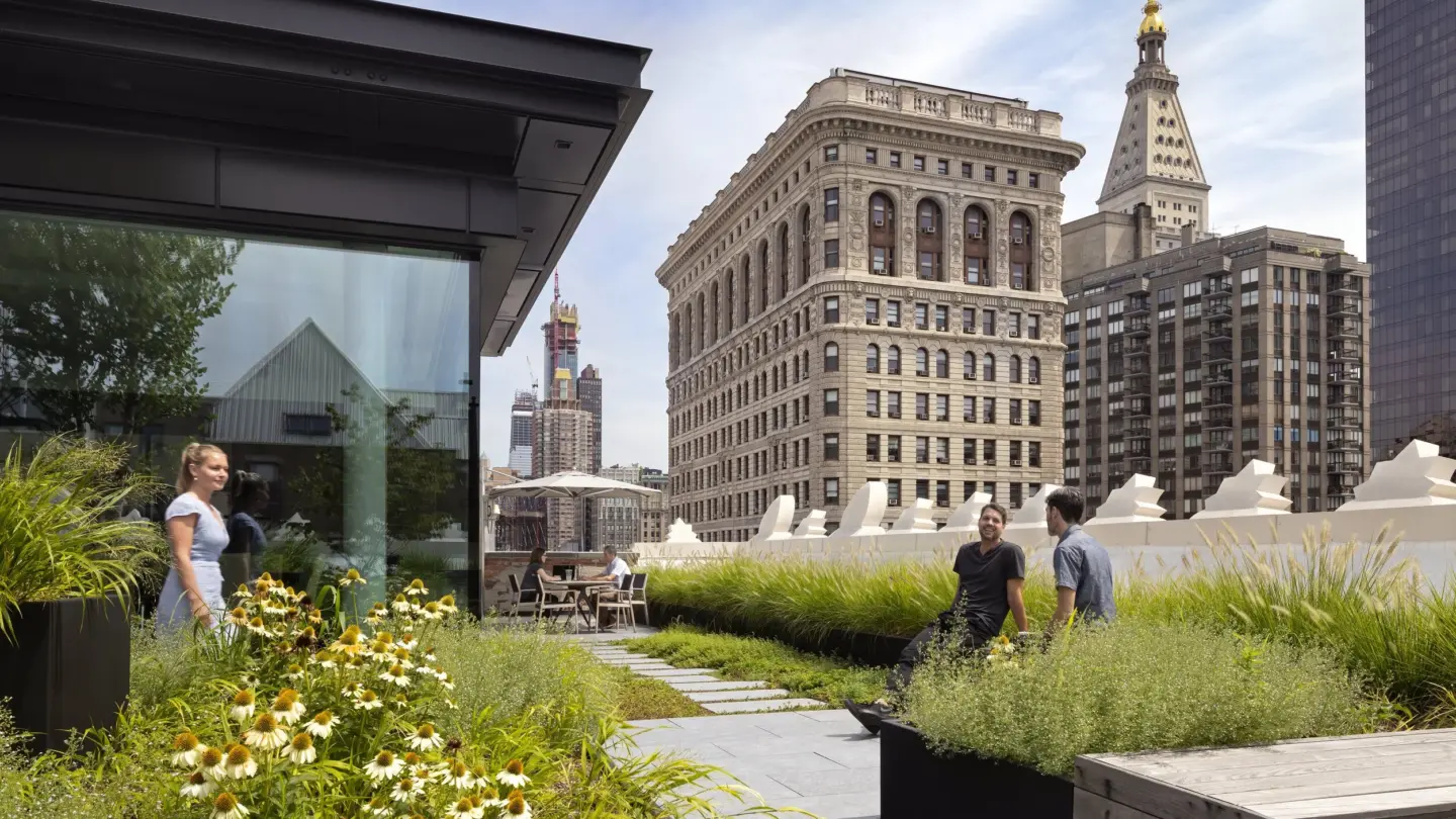 Three people walk and talk on a rooftop garden with greenery and flowers, surrounded by tall historic buildings under a blue sky.