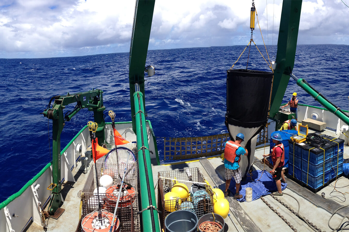 Scientists on a research ships deck handle oceanographic equipment and supplies under a blue sky, with the deep blue ocean in the background.