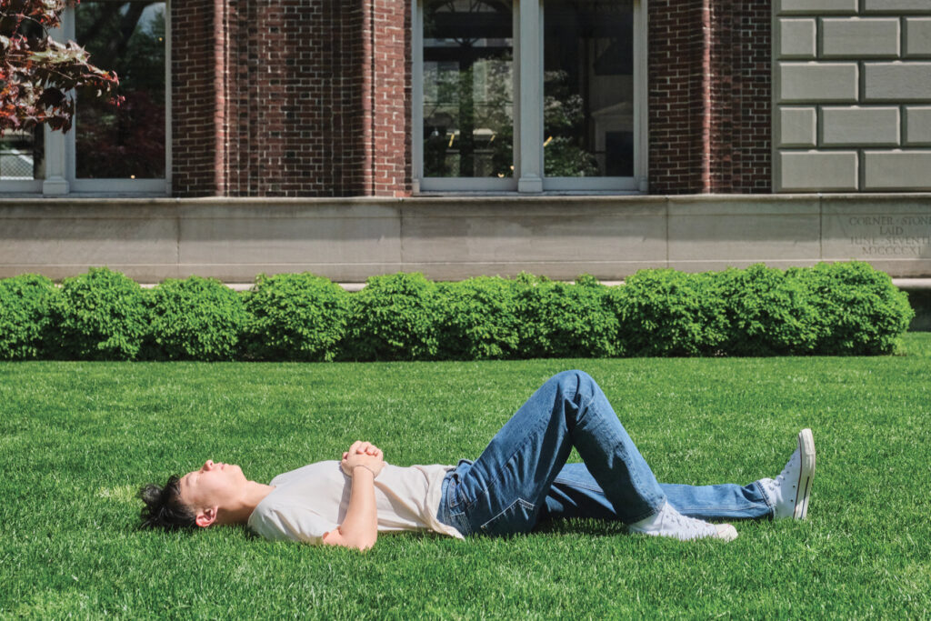 A person in a white shirt and blue jeans lies on their back on green grass in front of a brick building, eyes closed, enjoying the sunlight.