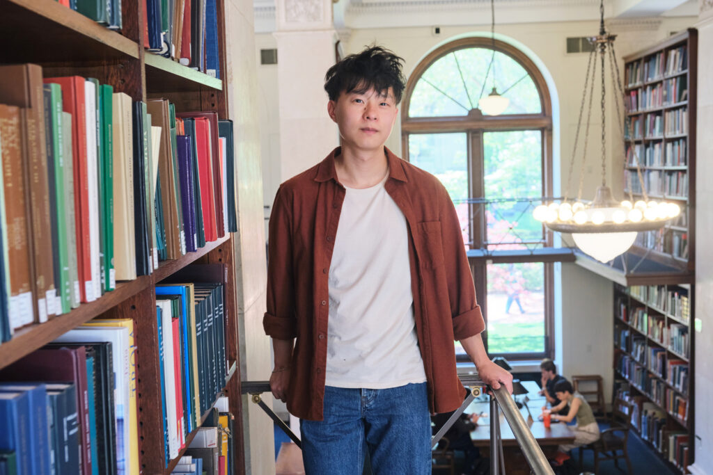 A person stands by bookshelves in a library, wearing a brown shirt over a white t-shirt and jeans. Large windows and people studying at tables are visible in the background.
