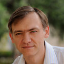 A man with short brown hair and a light complexion is wearing a white collared shirt, looking at the camera with a slight smile, with a blurred green outdoor background.