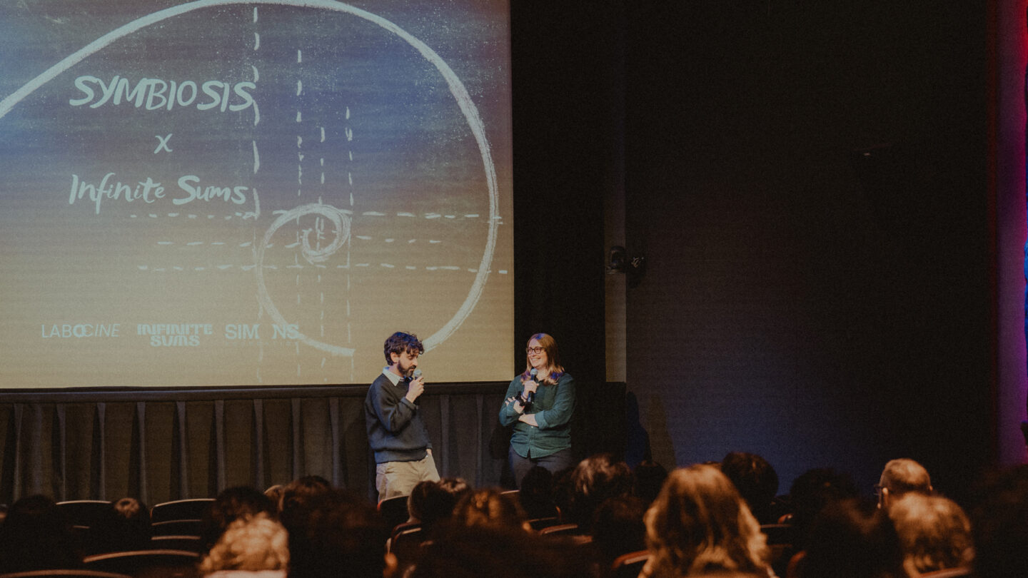 A photo of Sonya Hansen and Diego Murillo at the Symbiosis film screening, standing in front of a screen that says Symbiosis x Infinite Sums.