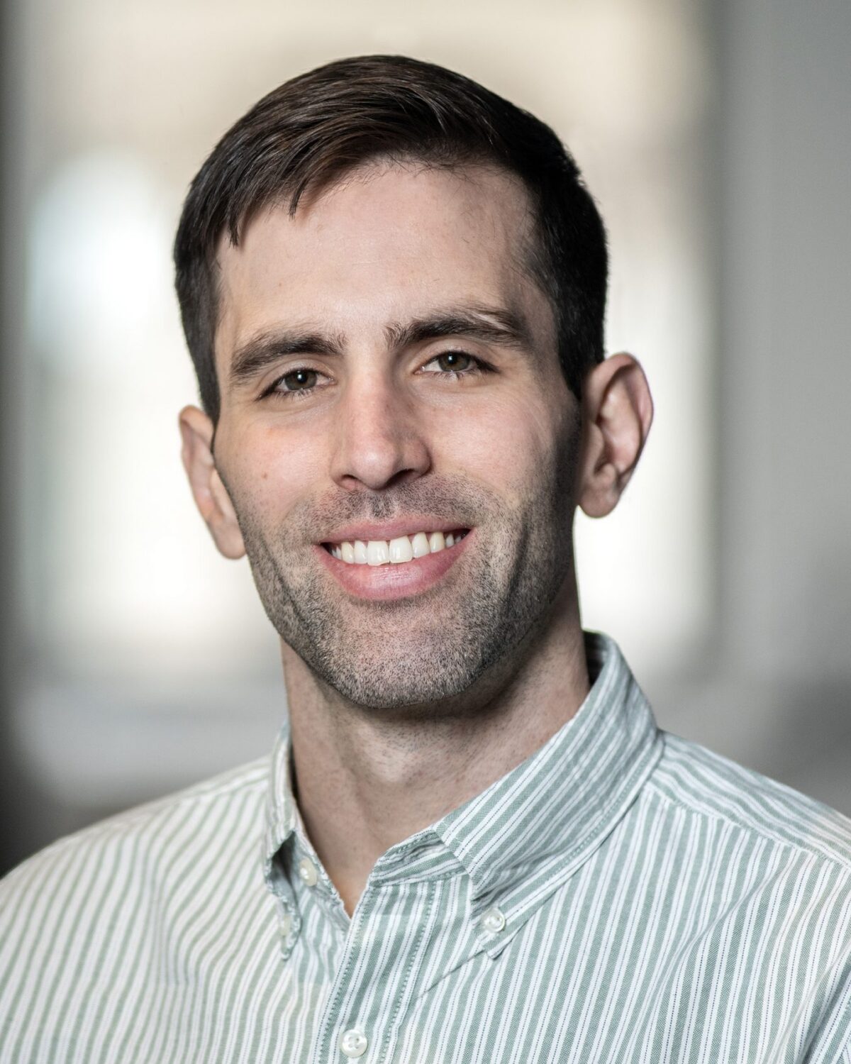 A man with short brown hair and a trimmed beard, wearing a light green and white striped button-up shirt, smiles in a softly lit indoor setting with blurred background.