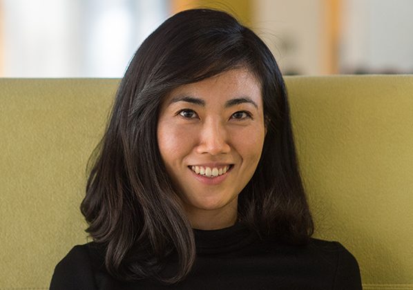 A woman with shoulder-length dark hair and a black top smiles while sitting on a light green chair, with a blurred background.
