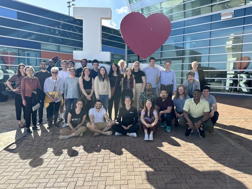 A group of people pose and smile in front of a large I ♥ sign outside a modern glass building on a sunny day.