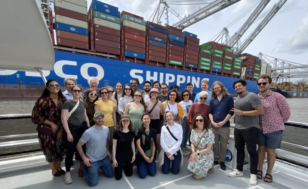 A group of people pose and smile on a boat with a large cargo ship and colorful shipping containers in the background, under a partly cloudy sky.
