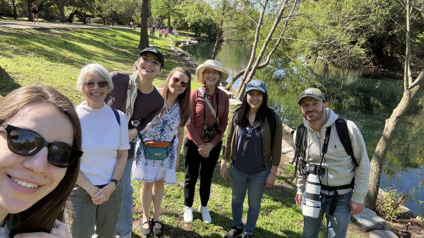 A group of seven people smiling and posing for a selfie outdoors on a sunny day by a pond, surrounded by green grass and trees.