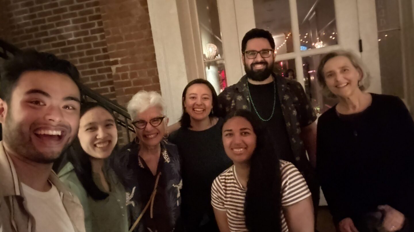 Seven people smiling and posing together indoors in front of a door with glass panes. The setting appears warm and friendly, with some exposed brick in the background and soft lighting.