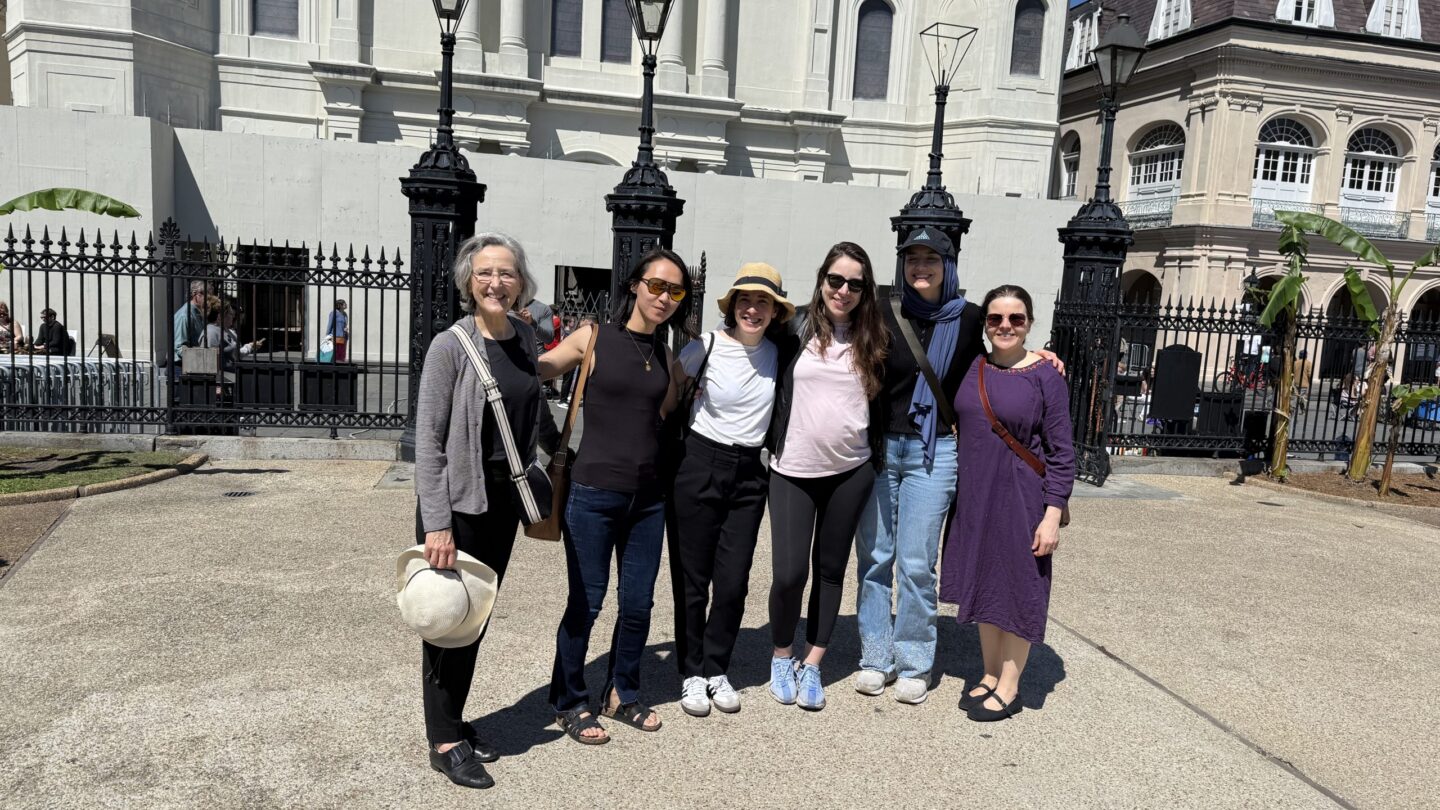 Six women stand together smiling in front of a historic white building and black iron fence on a sunny day. They are casually dressed and appear to be enjoying a group outing.