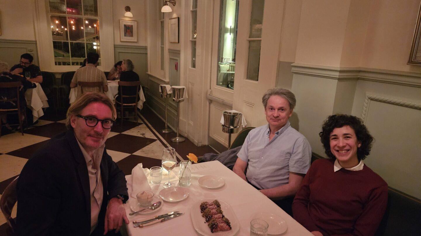 Three people sit at a restaurant table with food and drinks, smiling at the camera. The restaurant has elegant lighting, checkered floors, and other diners in the background.