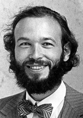 A man with a full beard and wavy hair smiles at the camera. He is wearing a suit, a striped bow tie, and a light-colored shirt. The image is black and white.