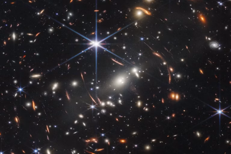 A dense field of distant galaxies with glowing stars, bright light flares, and scattered galaxies of various shapes and colors against the blackness of space.