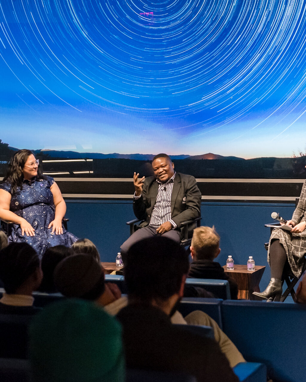 Three people sit on stage having a panel discussion in front of an audience, with a screen behind them displaying a night sky with star trails. Two women and one man are featured; plants decorate the stage.