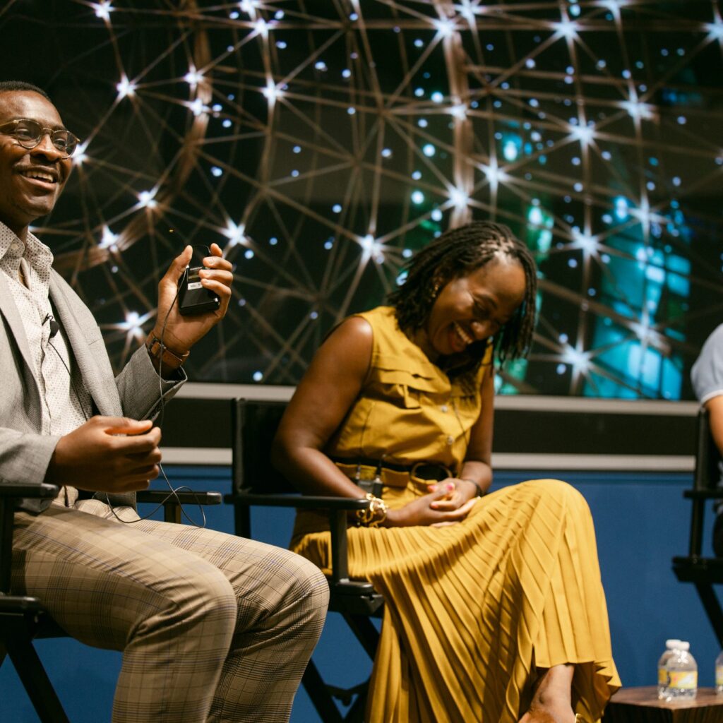Three people sit onstage, laughing and talking. The man on the left holds a microphone, the woman in the center wears a yellow dress, and the man on the right smiles. A backdrop of glowing lights is behind them.