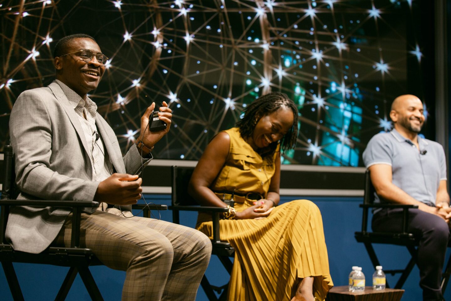 Three people sit onstage, laughing and talking. The man on the left holds a microphone, the woman in the center wears a yellow dress, and the man on the right smiles. A backdrop of glowing lights is behind them.