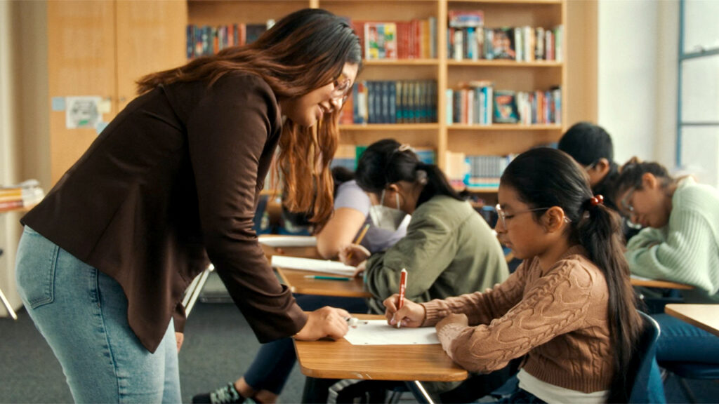 A teacher leans over to help a student seated at a desk in a classroom. Other students are writing or reading at their desks. Bookshelves are visible in the background.