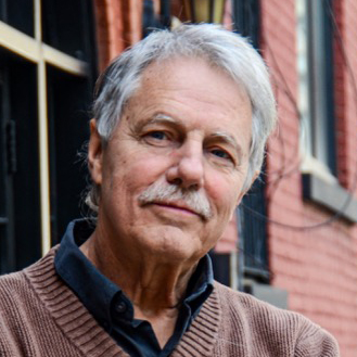 An older man with gray hair and a mustache, wearing a brown sweater over a dark shirt, stands in front of a brick building, looking calmly at the camera.