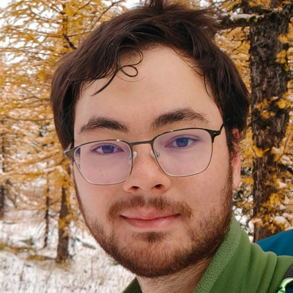 A person with glasses, dark hair, and a short beard smiles outdoors. Yellow autumn trees and a light layer of snow are visible in the background.