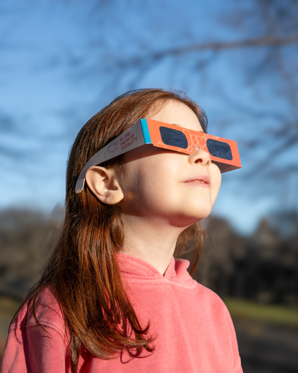 A young girl in a pink hoodie wears solar eclipse glasses and looks up at the sky in a sunlit park with bare trees and blurred buildings in the background.